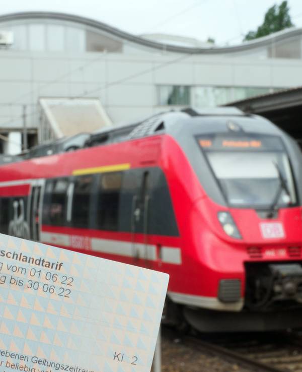 Die Hand eines Mannes hält ein Neun-Euro-Ticket. Auf einem Bahnsteig wartet dabei ein Regionalzug der Deutschen Bahn auf die Abfahrt.