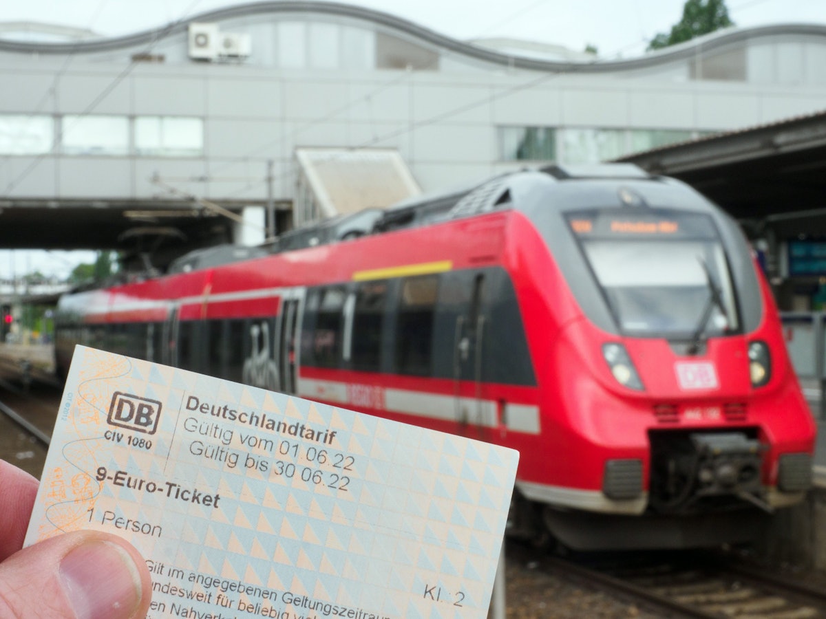 Die Hand eines Mannes hält ein Neun-Euro-Ticket. Auf einem Bahnsteig wartet dabei ein Regionalzug der Deutschen Bahn auf die Abfahrt.