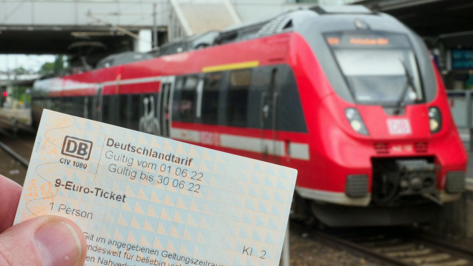 Die Hand eines Mannes hält ein Neun-Euro-Ticket. Auf einem Bahnsteig wartet dabei ein Regionalzug der Deutschen Bahn auf die Abfahrt.