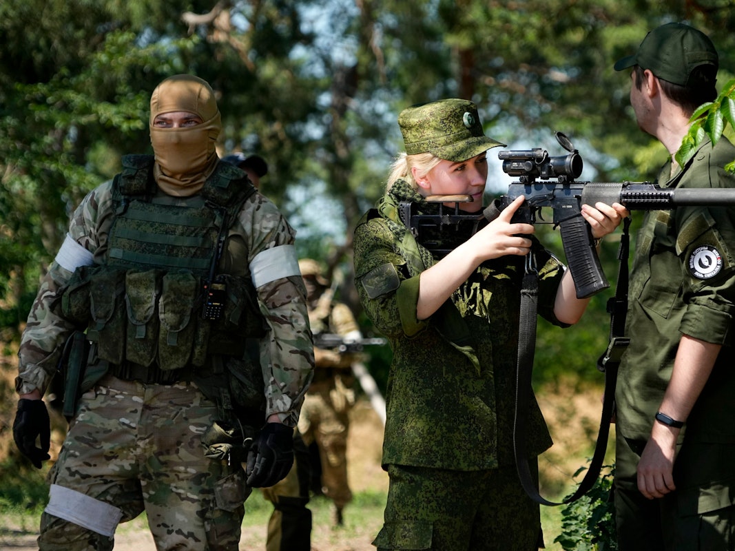 Eine Soldatin der russischen Armee blickt durch ein Scharfschützenzielfernrohr, während sie und andere Soldaten und Soldatinnen eine Gruppe ausländischer Journalisten und Journalistinnen bewachen.