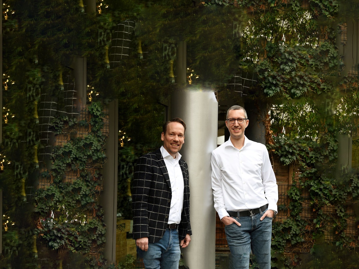 Zwei Männer lehnen an einer Säule und lächeln in die Kamera.