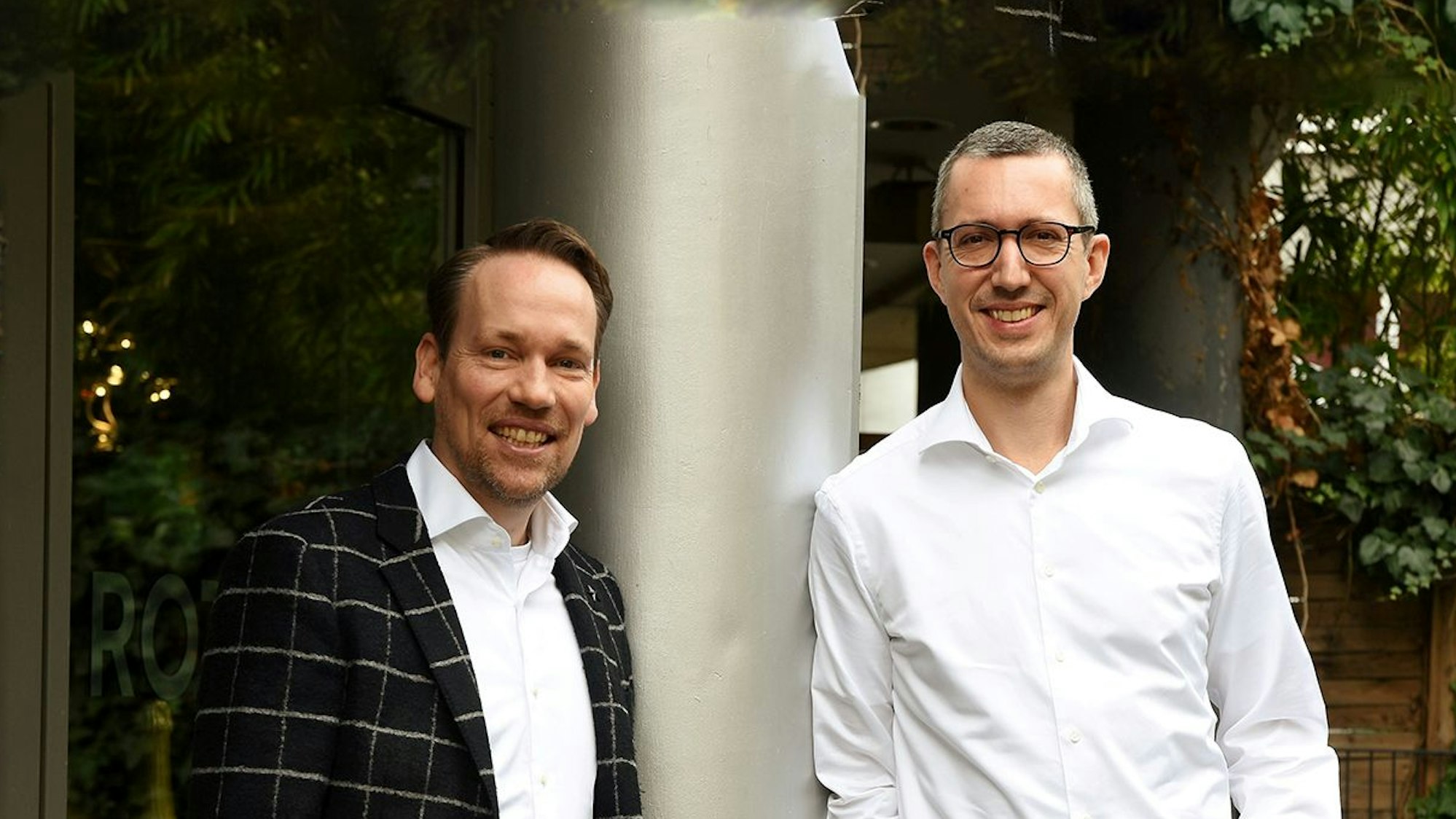 Zwei Männer lehnen an einer Säule und lächeln in die Kamera.