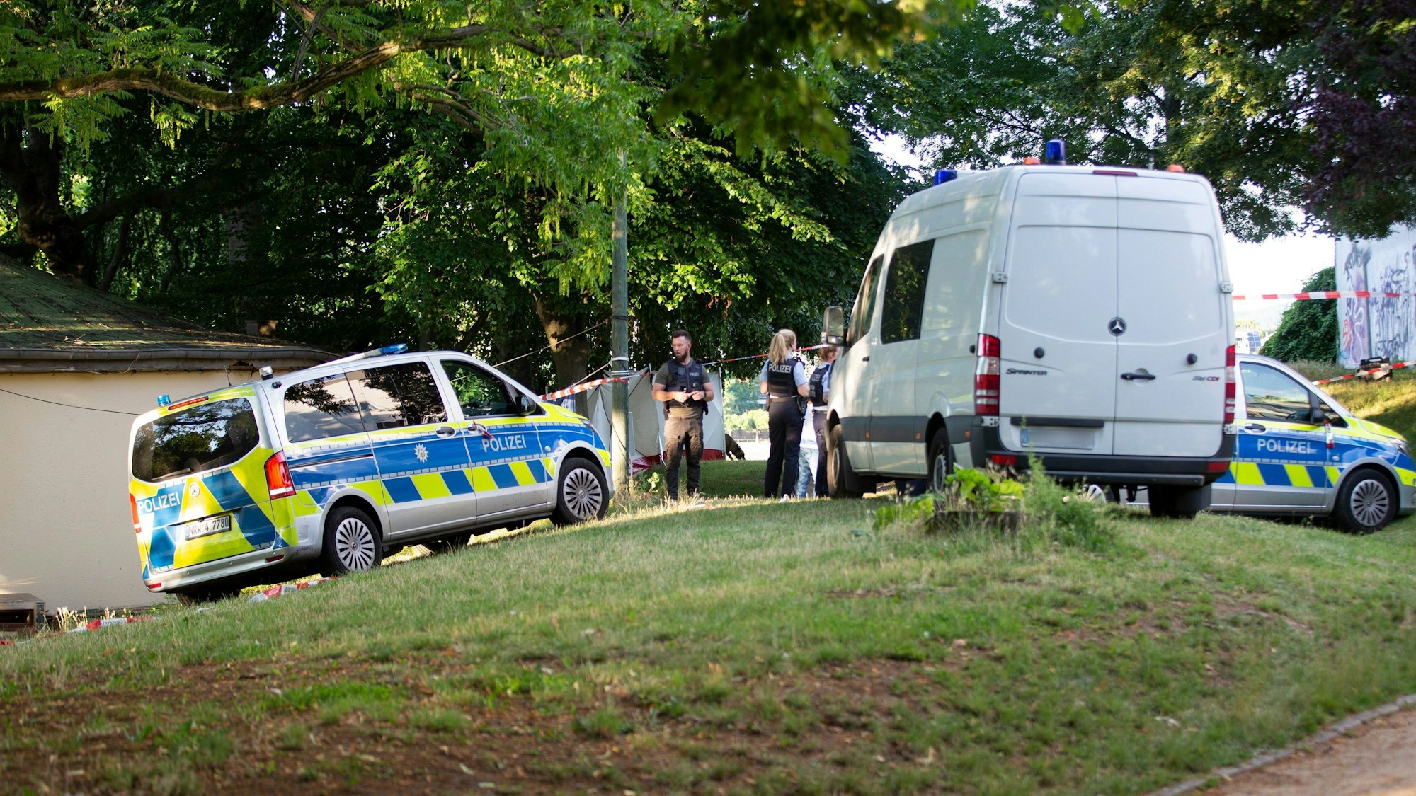 Fahrzeuge von Polizei und Spurensicherung parken auf einer Grünfläche vor einer Polizeiabsperrung.