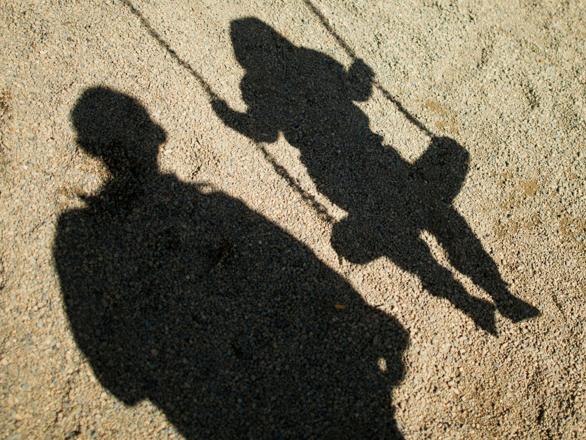 Der Schatten von einem Mann und einem schaukelnden Kind fallen auf Sand auf einem Spielplatz.