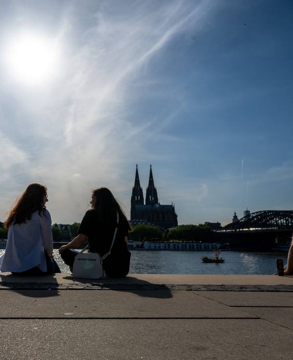 Zwei Frauen unterhalten sich am Rheinboulevard mit Blick zum Kölner Dom bei Sonnenschein.