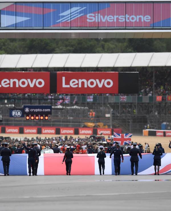 Auf der Start- und Zielgeraden hissen mehrere Personen die Flagge Großbritanniens – im Hintergrund bereiten sich die Teams auf das anstehende Rennen vor.