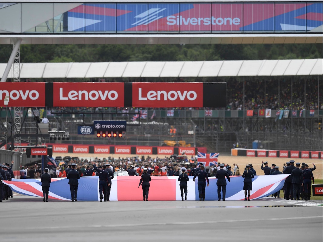 Auf der Start- und Zielgeraden hissen mehrere Personen die Flagge Großbritanniens – im Hintergrund bereiten sich die Teams auf das anstehende Rennen vor.