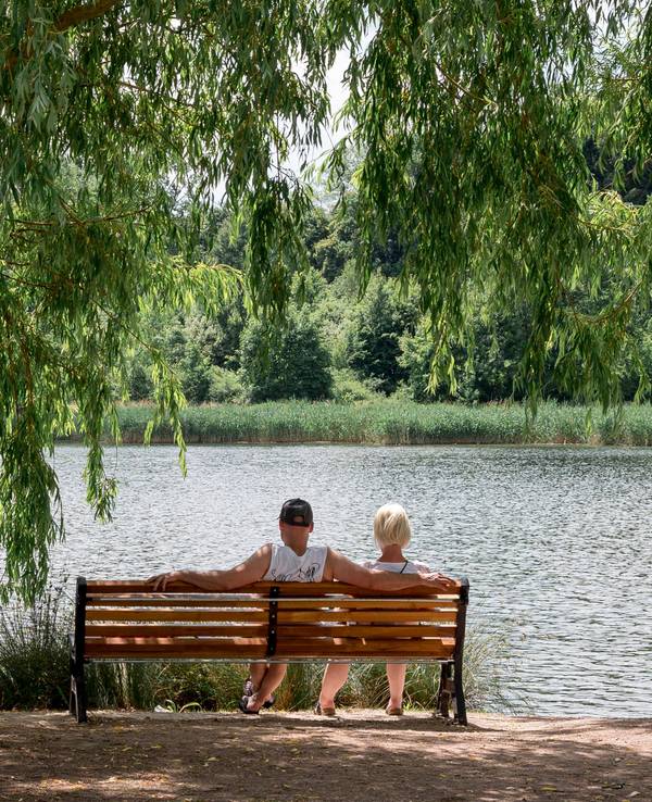 Ein Paar sitzt auf einer Bank in der Sonne an einem See.