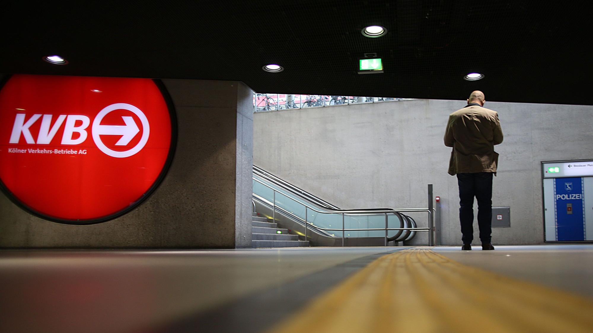Ein Mann steht in einer leeren U-Bahn Station und blickt auf sein Smartphone, neben ihm hängt ein KVB-Logo.