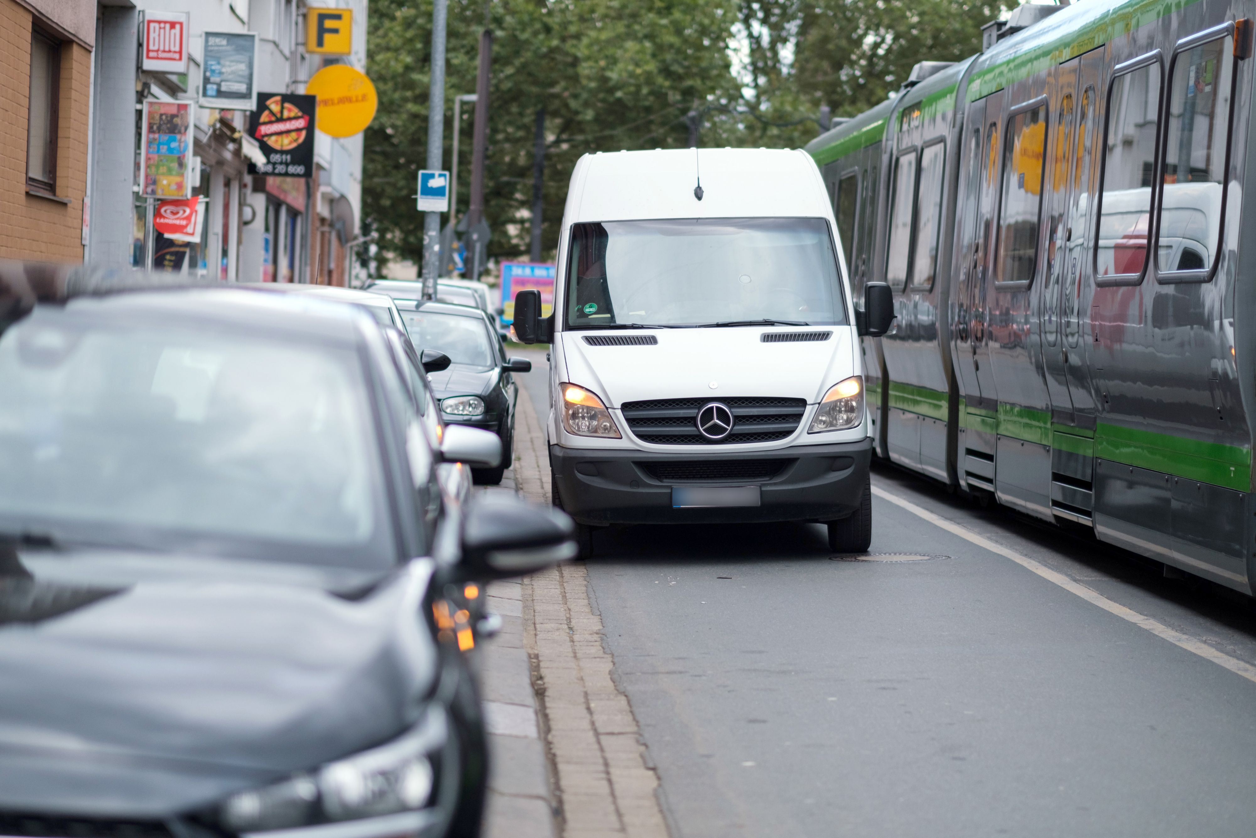 Ein Lieferwagen steht auf einer Straße neben parkenden Autos, daneben fährt eine Straßenbahn entlang.