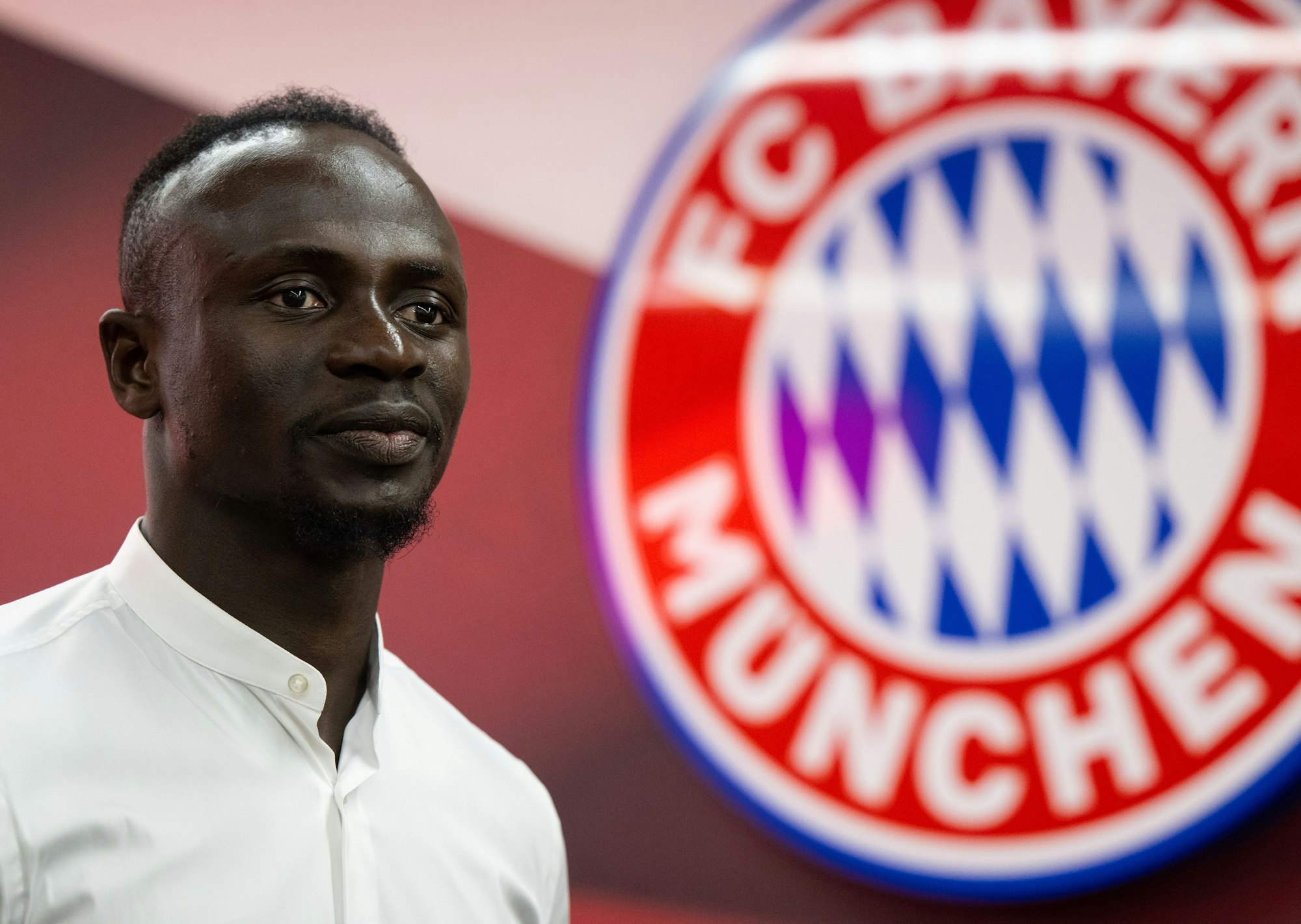 Sadio Mané posiert im weißen Hemd, hinter ihm ist das Wappen des FC Bayern zu sehen.