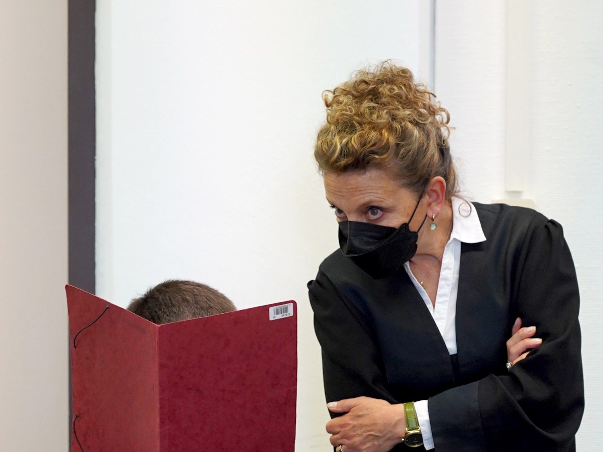 Ein Mann hält sich in einem Gerichtssaal eine rote Mappe vor das Gesicht.