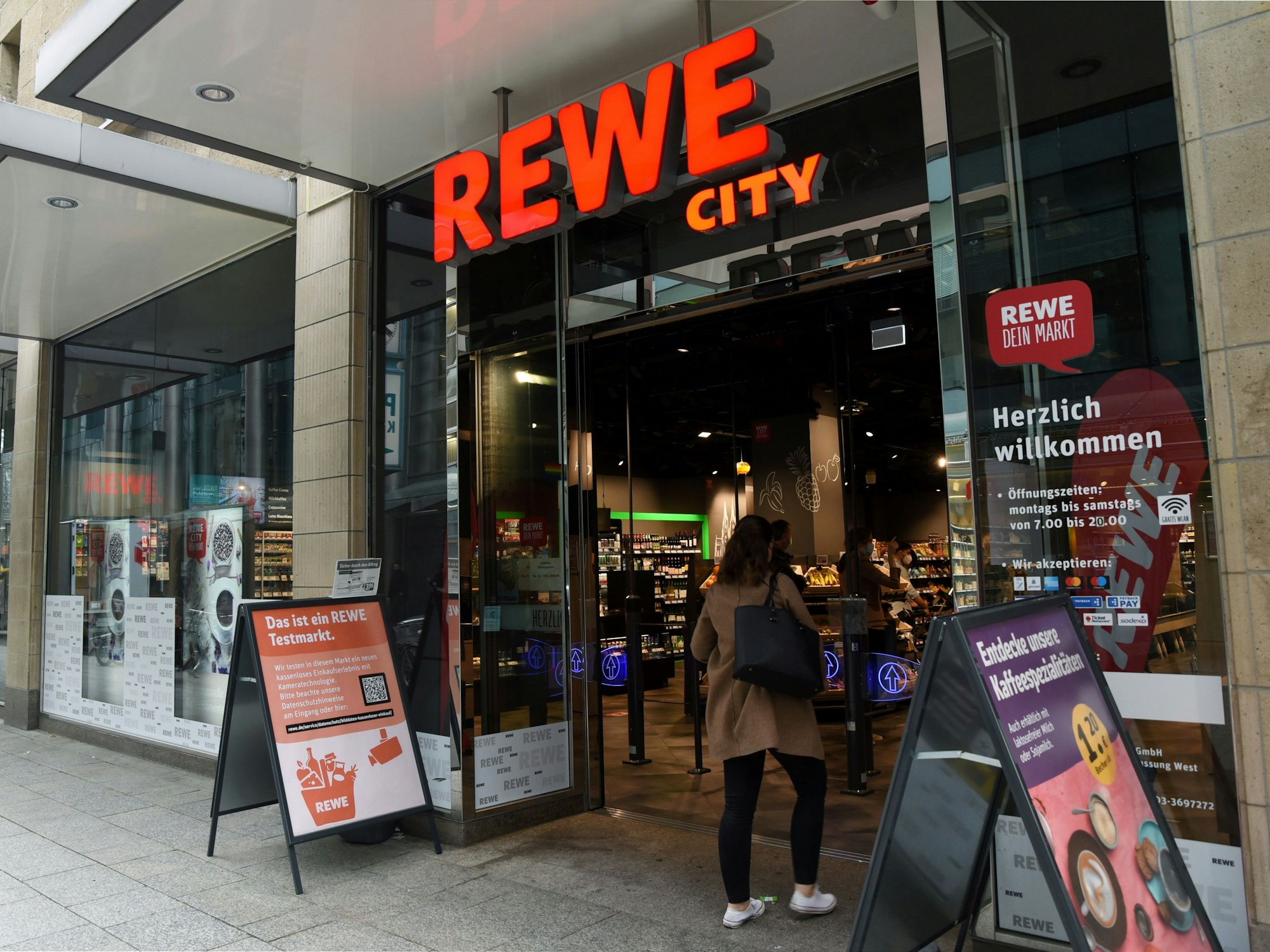 Eine Frau geht durch den Eingang einer Rewe City-Filiale in den Supermarkt.