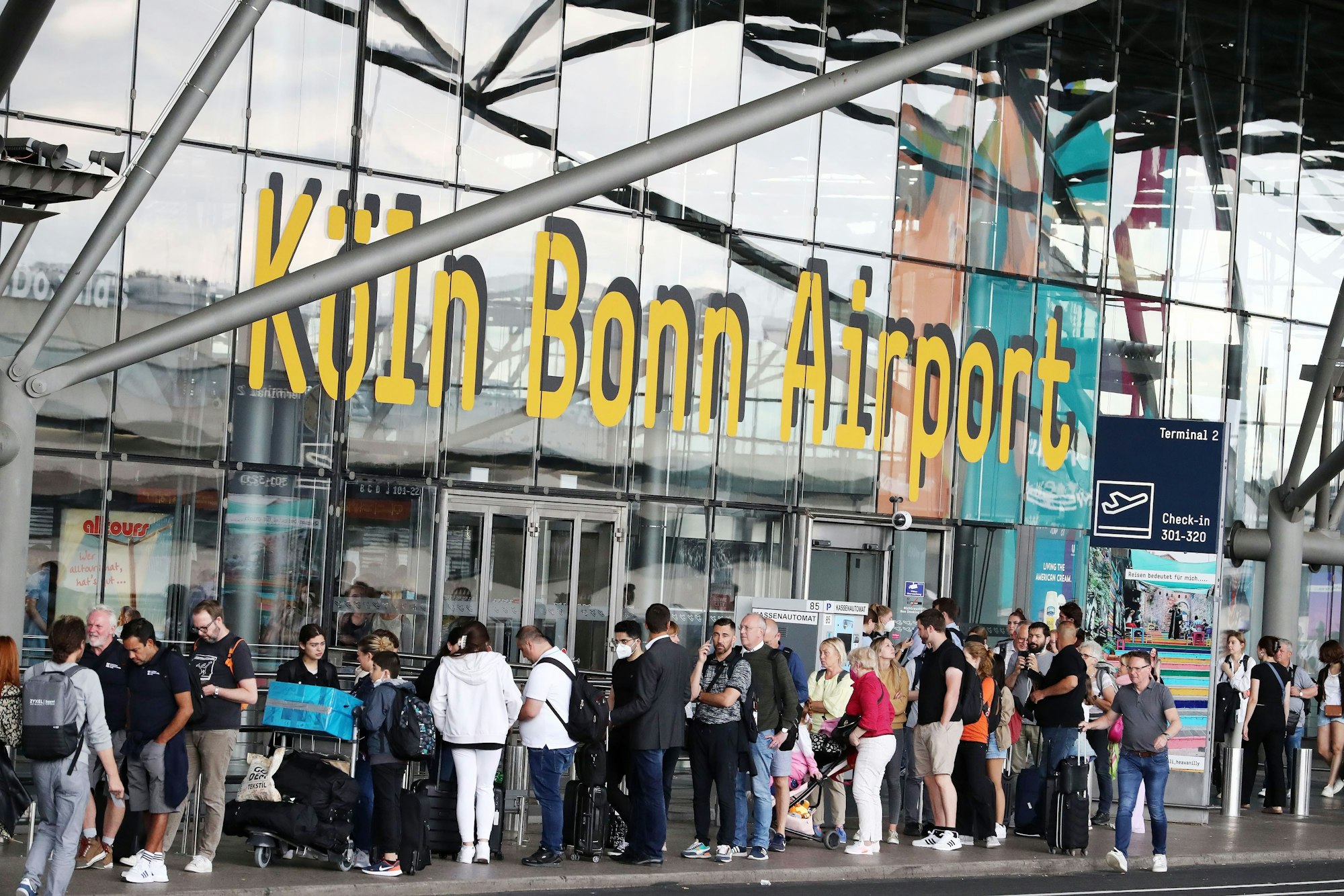 Fluggäste in der Warteschlange, die bis vor das Flughafengebäude reicht.