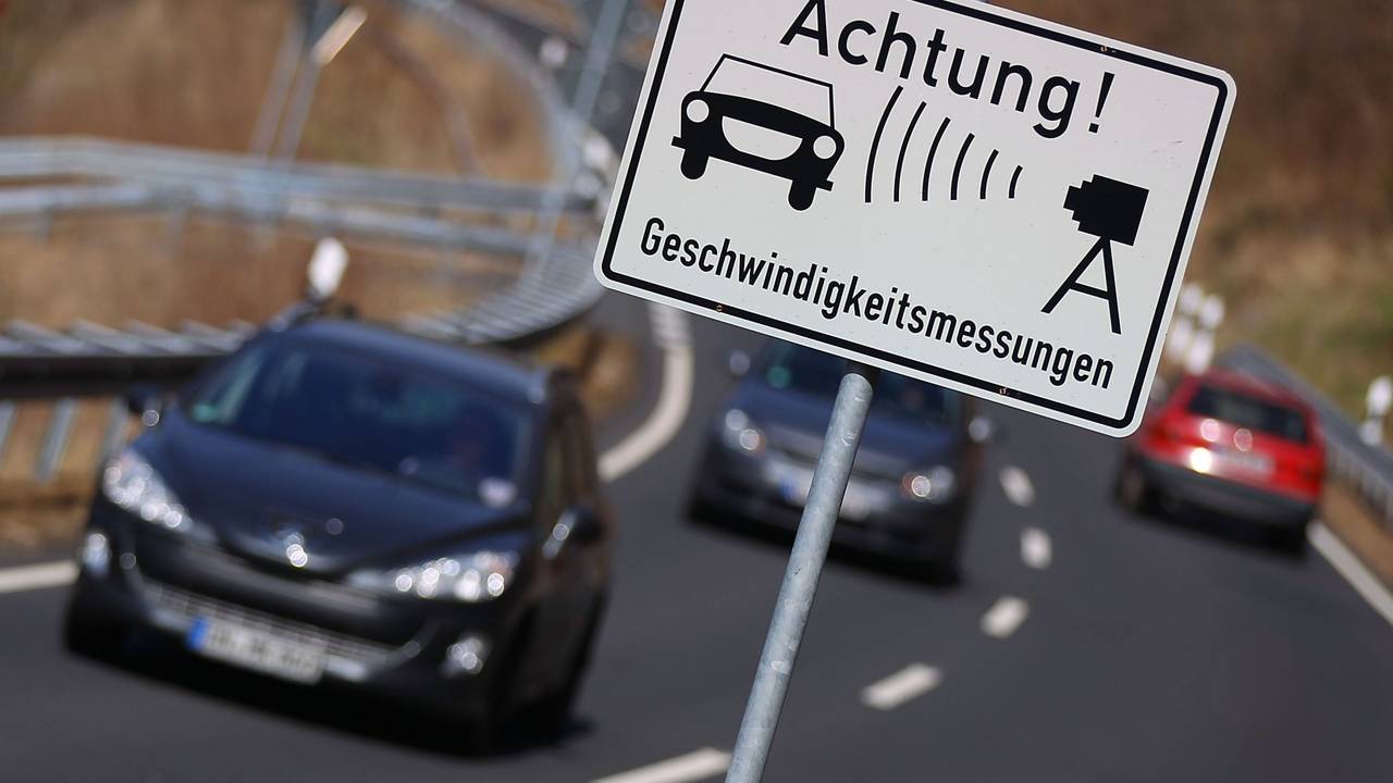Autos fahren auf einer Landstraße an einem Warnschild für Geschwindigkeitsmessungen vorbei.