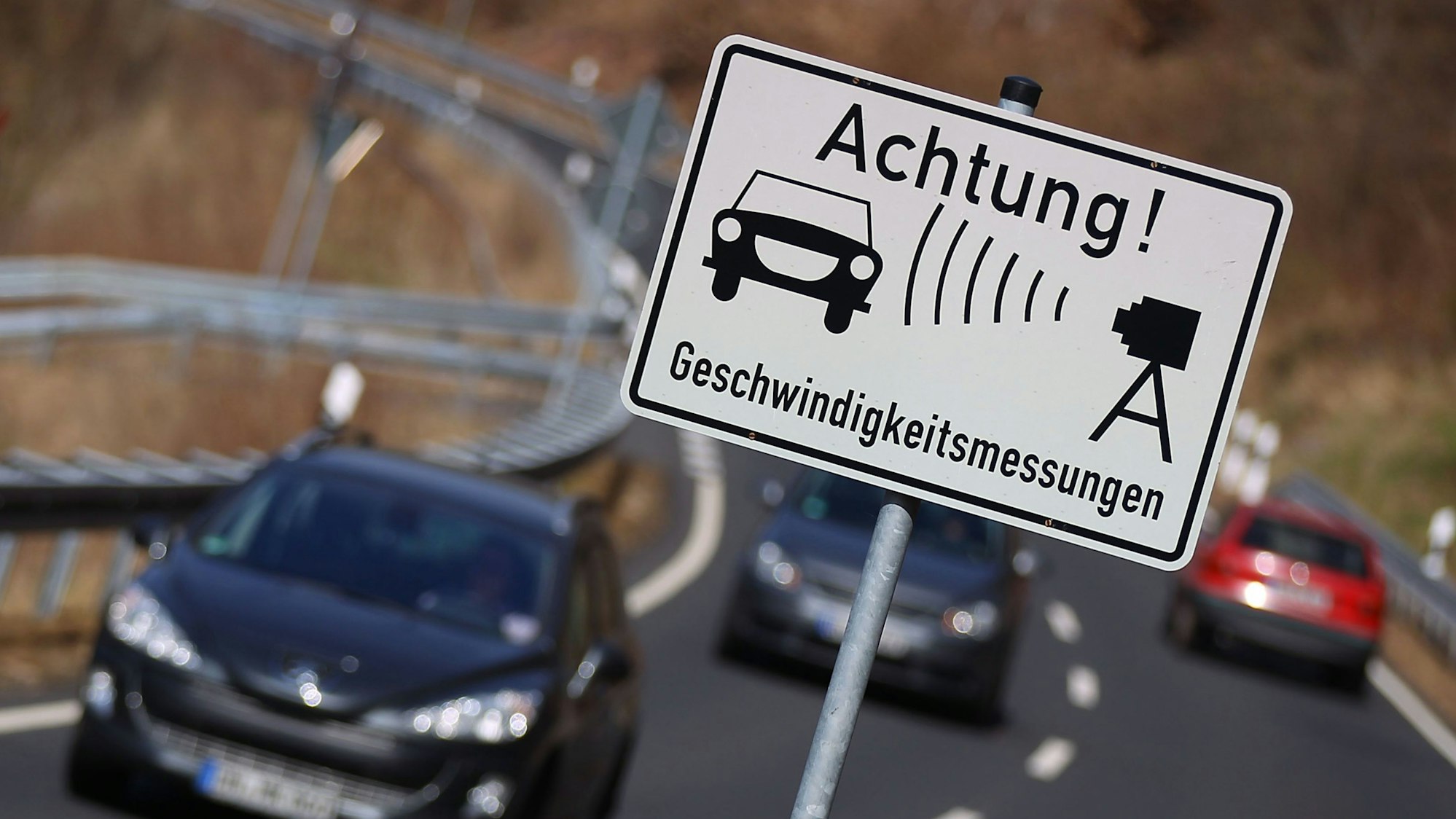 Autos fahren auf einer Landstraße an einem Warnschild für Geschwindigkeitsmessungen vorbei.