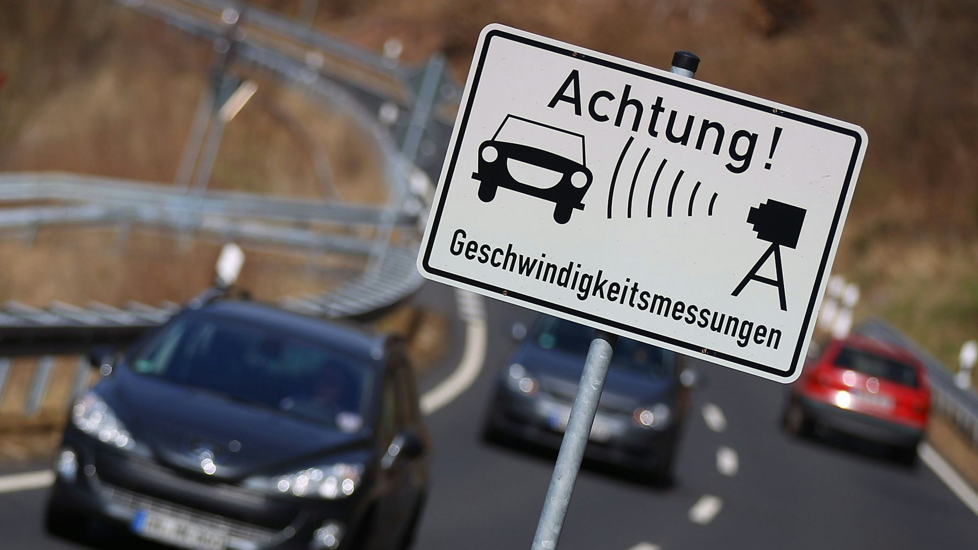 Autos fahren auf einer Landstraße an einem Warnschild für Geschwindigkeitsmessungen vorbei.