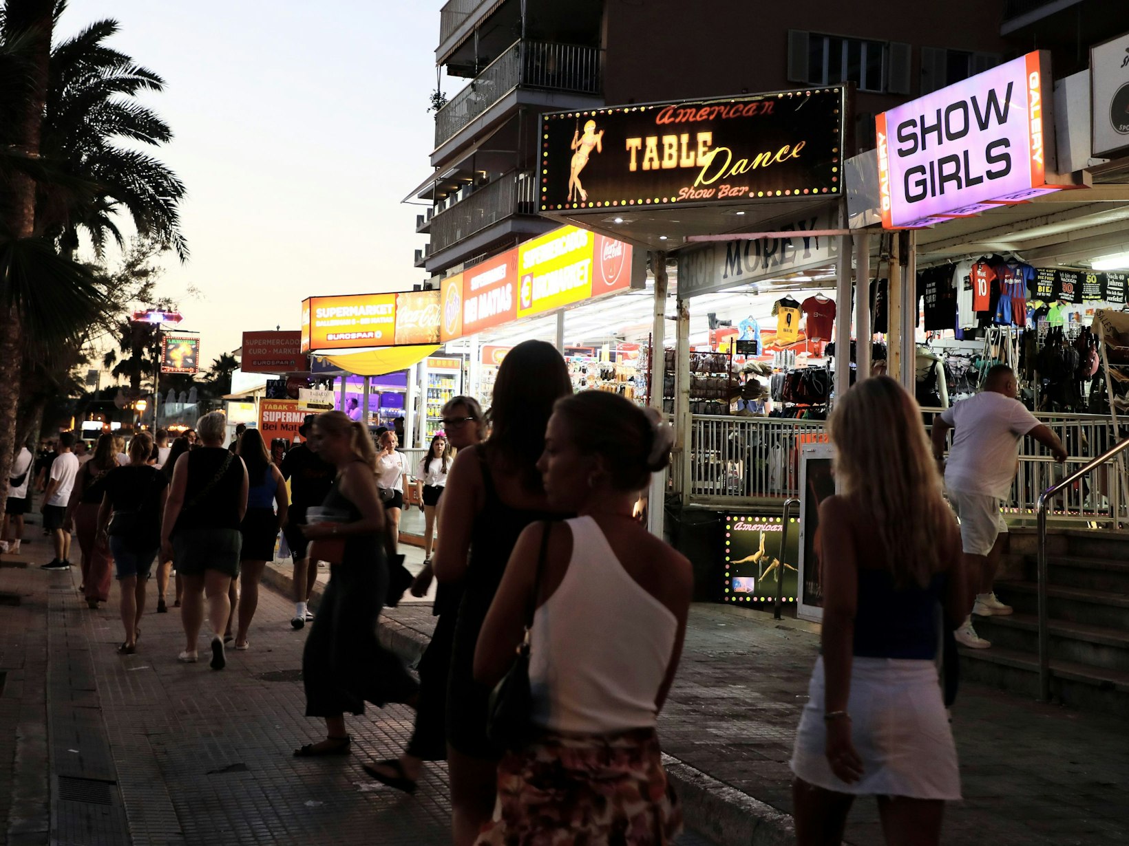 Menschen spazieren auf der Strandpromenade von Arenal auf Mallorca an Show-Bars vorbei.