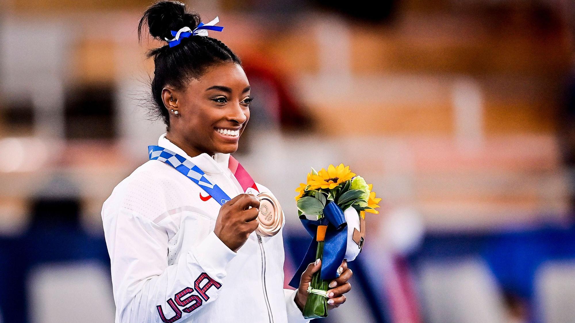Die Turnerin Simone Biles posiert mit Bronzemedaille und Blumenstrauß für die Kamera.