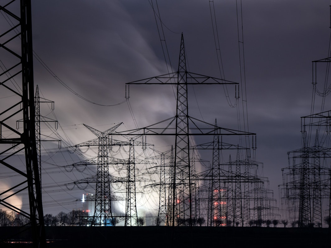 Hinter zahlreichen Strommasten zeichnet sich heller Dampf über dem Braunkohlekraftwerk Niederaußem vor dem noch dunklen Morgenhimmel ab.