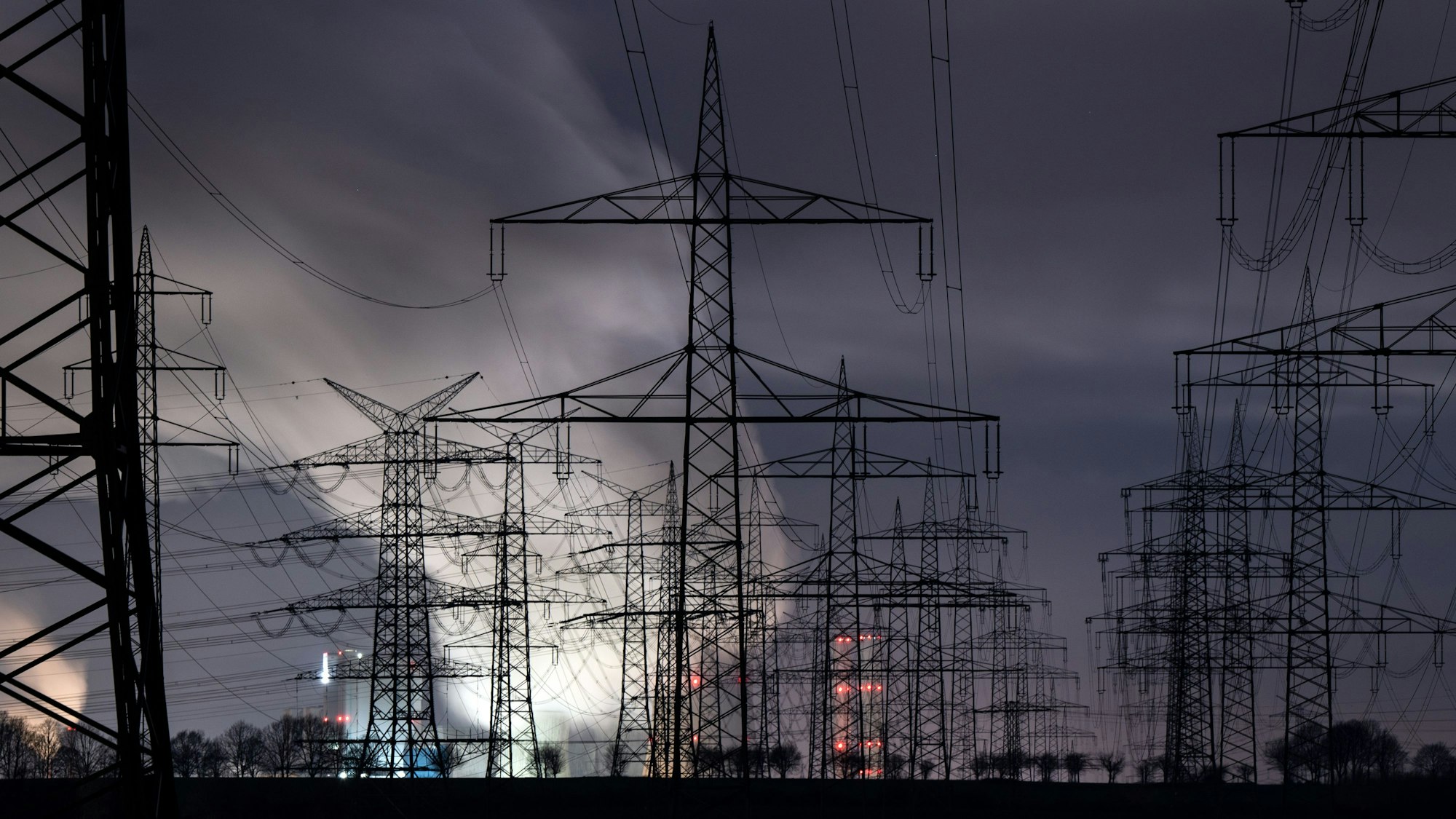 Hinter zahlreichen Strommasten zeichnet sich heller Dampf über dem Braunkohlekraftwerk Niederaußem vor dem noch dunklen Morgenhimmel ab.