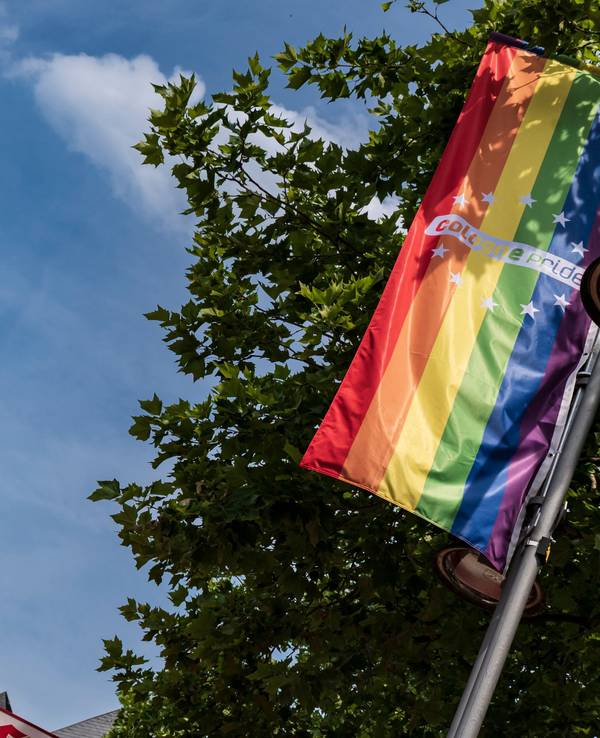 Eine LGBTIQ+-Flagge hängt an einer Straßenlaterne, im Hintergrund die Spitzen des Kölner Doms.