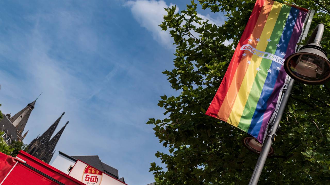Eine LGBTIQ+-Flagge hängt an einer Straßenlaterne, im Hintergrund die Spitzen des Kölner Doms.