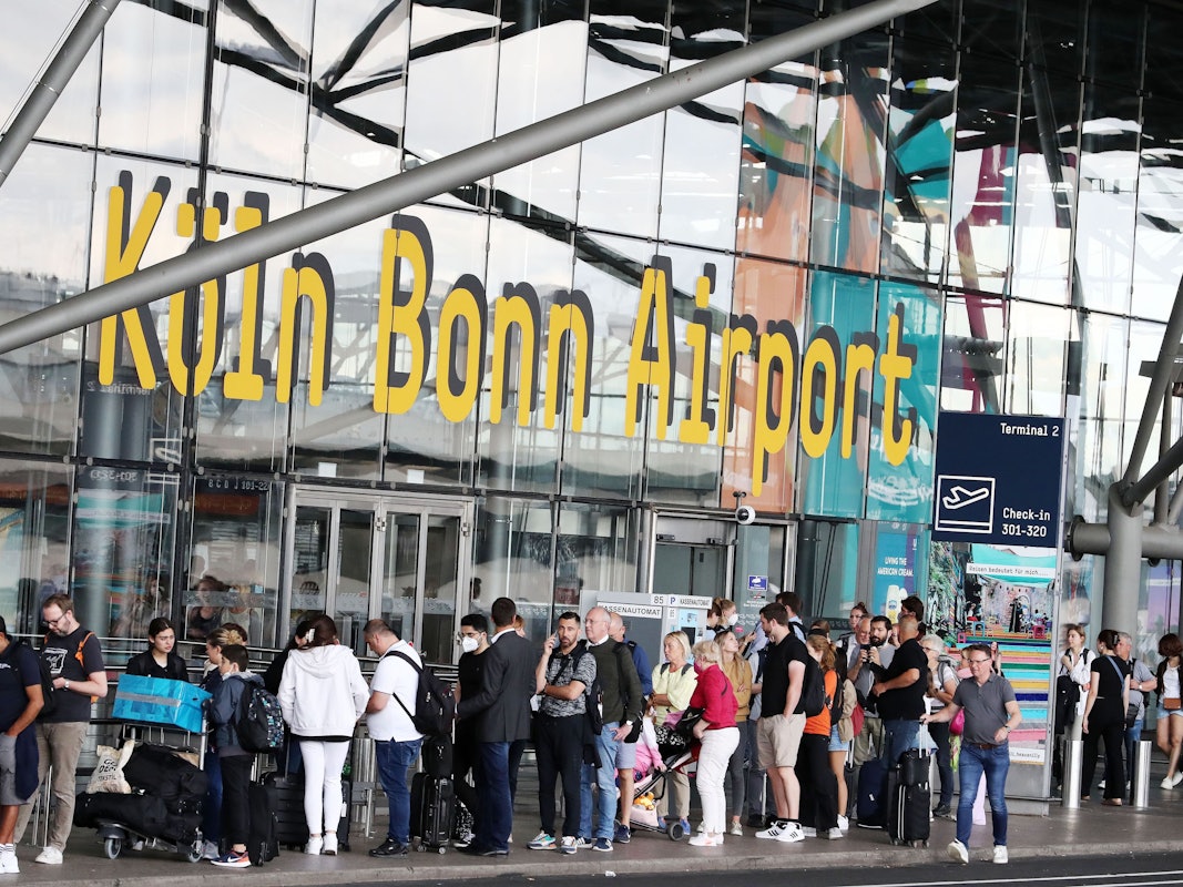 Fluggäste stehen vor einem Gebäude mit der Aufschrift "Köln Bonn Airport" in der Warteschlange.