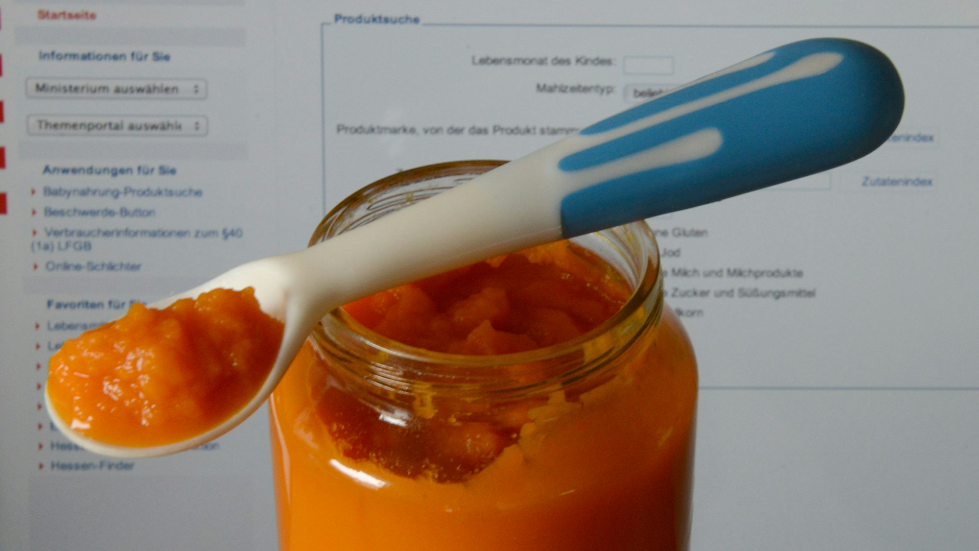 Ein Glas mit Karottenbrei für Babys steht vor einem Laptop-Monitor mit der geöffneten Seite einer Online-Datenbank zur Babynahrungs-Produktsuche.