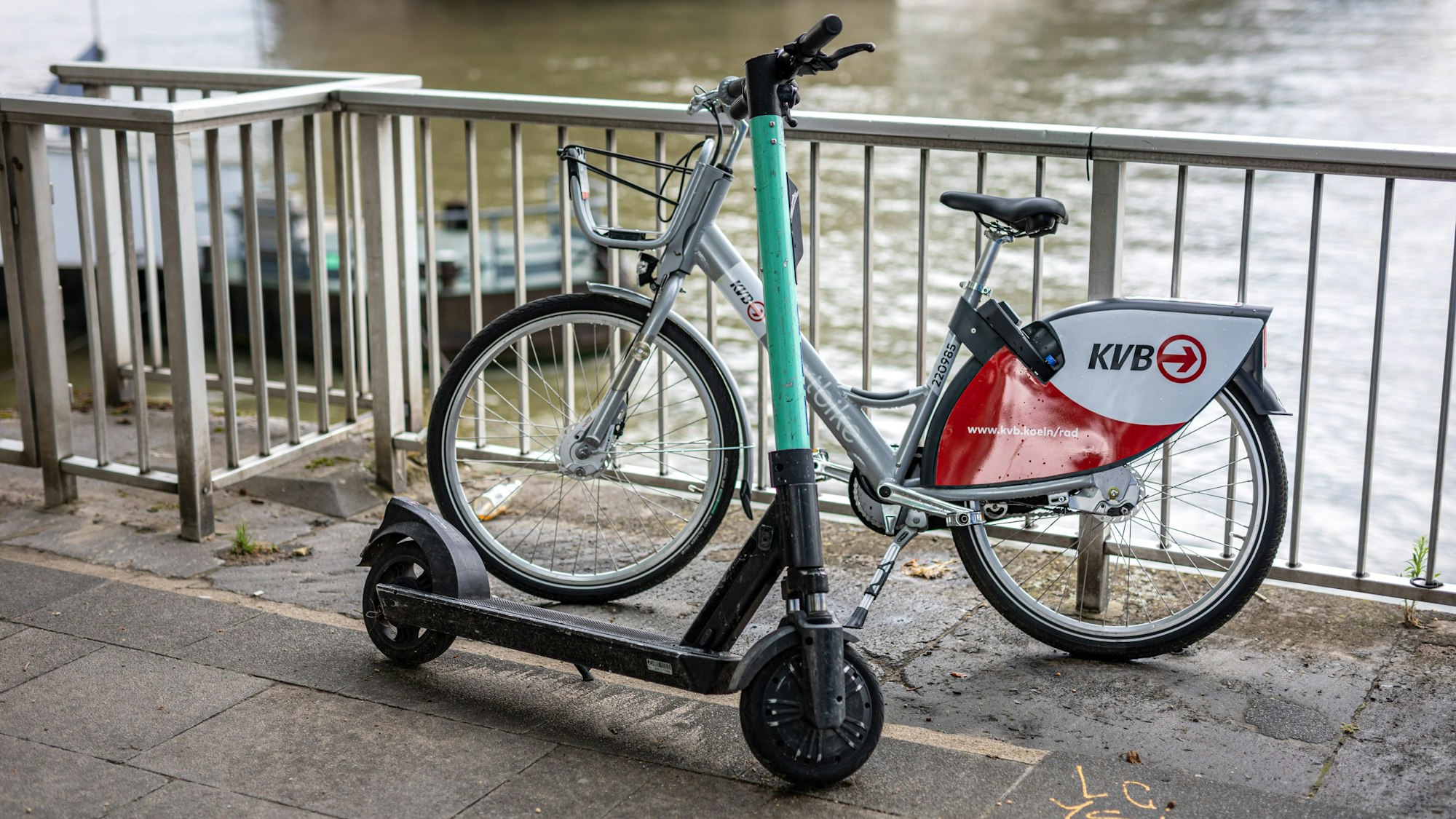 Ein E-Scooter und ein Leihfahrrad der Kölner Verkehrsbetriebe (KVB) stehen am Rheinufer am Geländer.