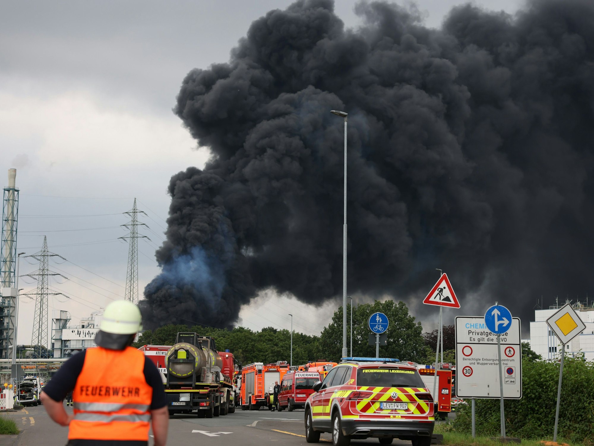 Einsatzfahrzeuge der Feuerwehr stehen unweit einer Zufahrt zum Chemiepark, über dem eine dunkle Rauchwolke aufsteigt. Am 27. Juli 2021 war es in Leverkusen zu einer verheerenden Explosion gekommen. Sieben Mitarbeiter starben, viele wurden verletzt.