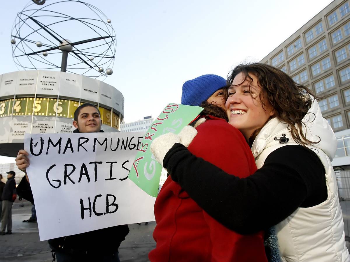 Menschen geben sich auf dem Alexanderplatz in Berlin eine Umarmung, ein weiterer hält ein Schild mit dem Text „Umarmung gratis“ hoch.