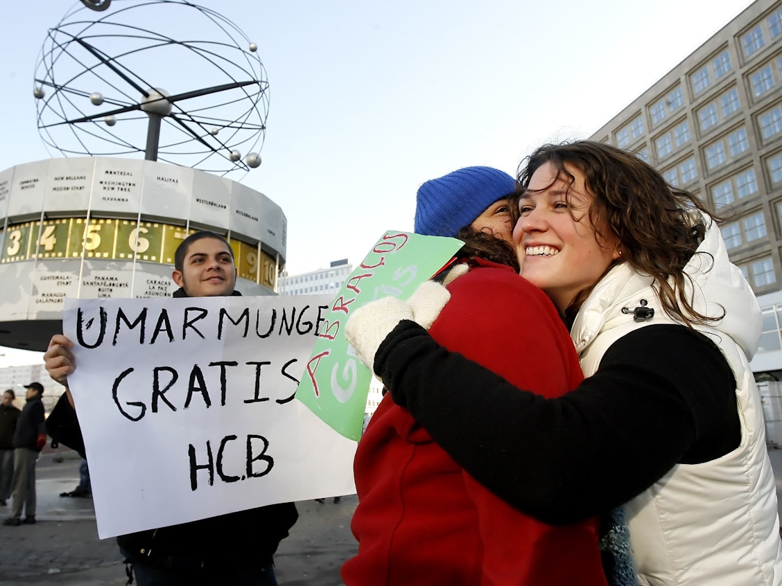 Menschen geben sich auf dem Alexanderplatz in Berlin eine Umarmung, ein weiterer hält ein Schild mit dem Text „Umarmung gratis“ hoch.