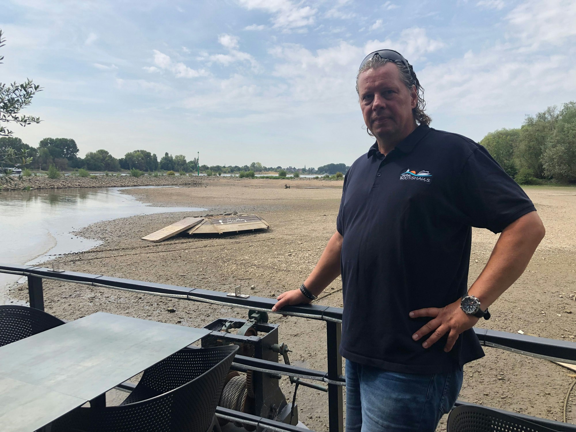 Jörg Blöck steht am Geländer des Außenbereiches des Restaurants, hinter ihm der trockene Grund des Rheins.