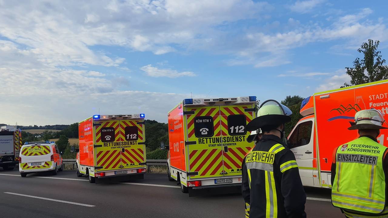 Zwei Feuerwehrmänner stehen mit dem Rücken zur Kamera vor ihren Einsatzwagen auf der Autobahn.