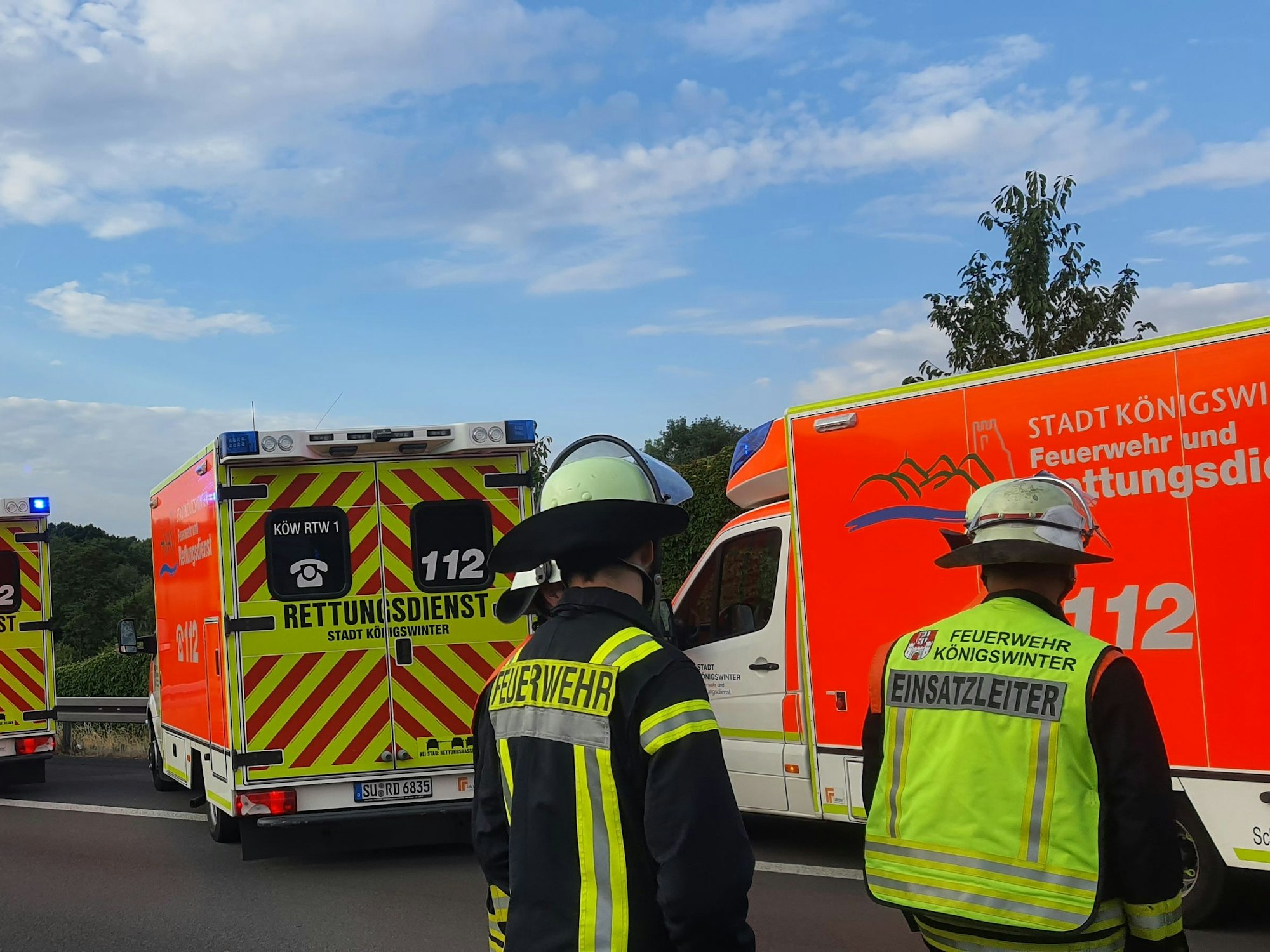 Zwei Feuerwehrmänner stehen mit dem Rücken zur Kamera vor ihren Einsatzwagen auf der Autobahn.