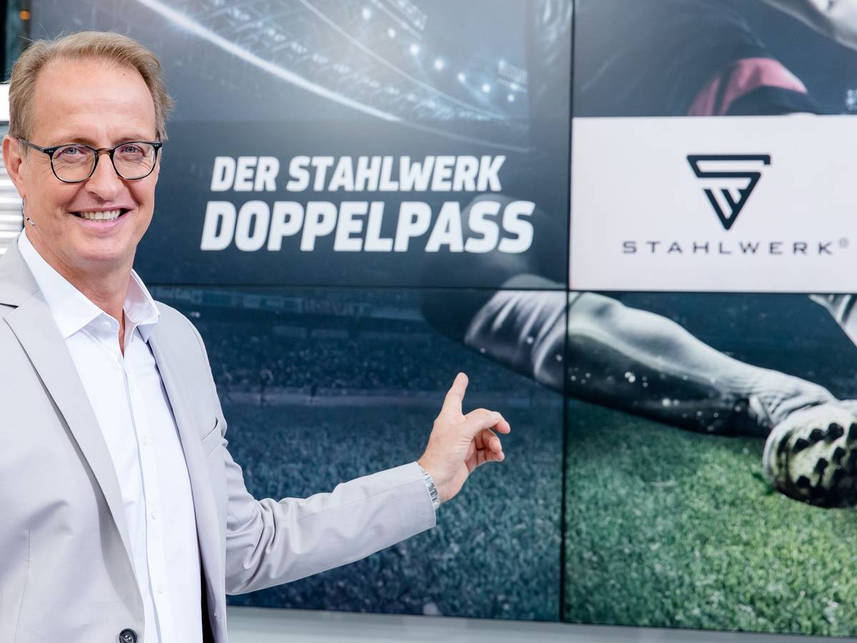 Florian König zeigt auf das Logo des Fußball-Talks Doppelpass. Er wird den Doppelpass auch in der Saison 2022/2023 moderieren.