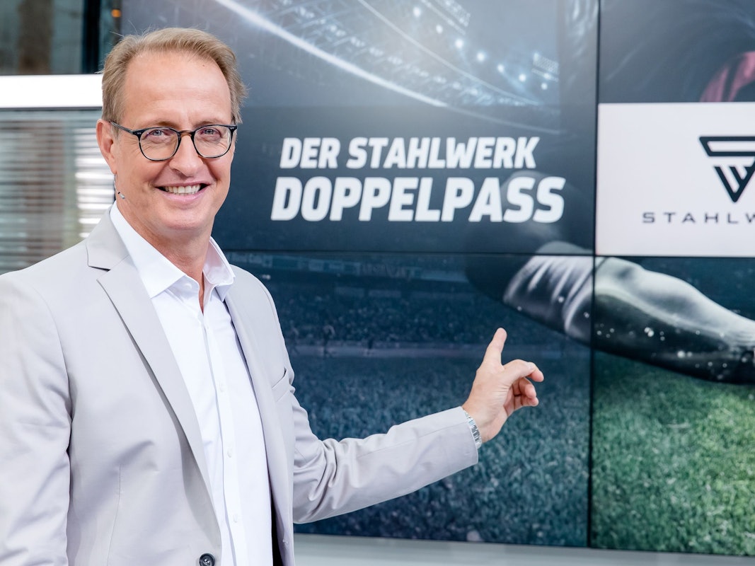 Florian König zeigt auf das Logo des Fußball-Talks Doppelpass. Er wird den Doppelpass auch in der Saison 2022/2023 moderieren.