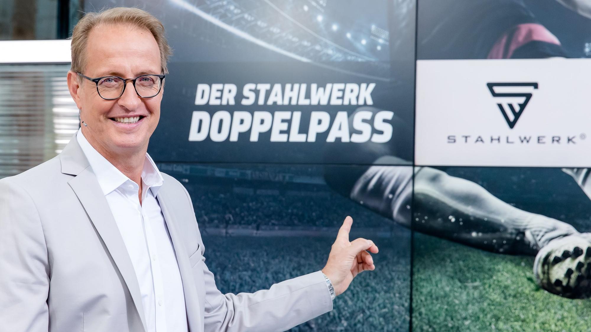 Florian König zeigt auf das Logo des Fußball-Talks Doppelpass. Er wird den Doppelpass auch in der Saison 2022/2023 moderieren.