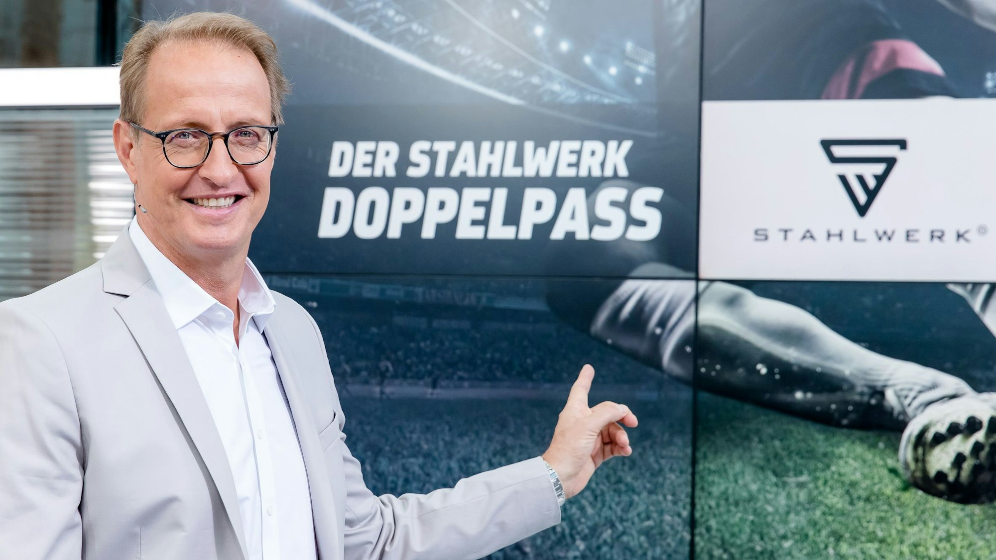 Florian König zeigt auf das Logo des Fußball-Talks Doppelpass. Er wird den Doppelpass auch in der Saison 2022/2023 moderieren.
