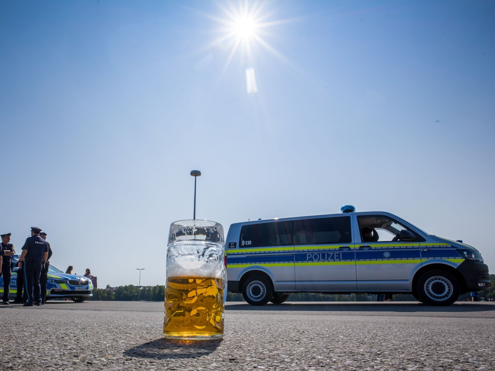 Ein verlassener, halb gefüllter Bierkrug auf dem Asphalt. Im Hintergrund stehen Polizisten neben Polizeifahrzeugen.