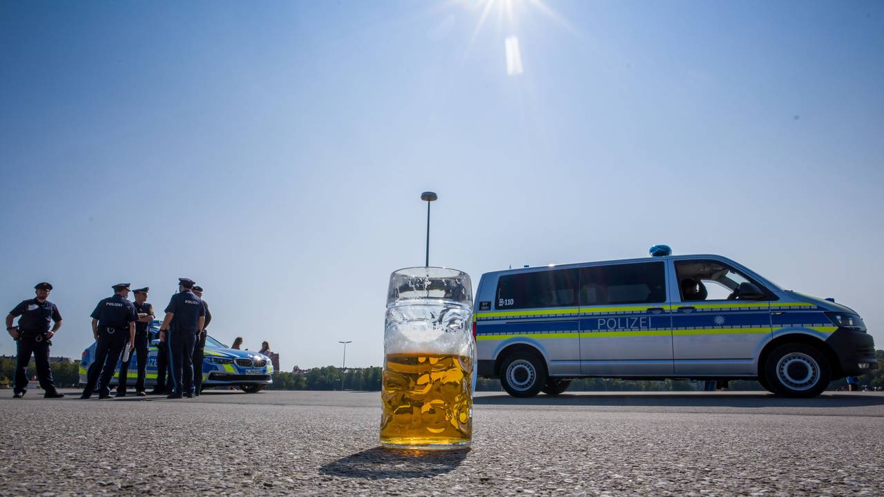 Ein verlassener, halb gefüllter Bierkrug auf dem Asphalt. Im Hintergrund stehen Polizisten neben Polizeifahrzeugen.