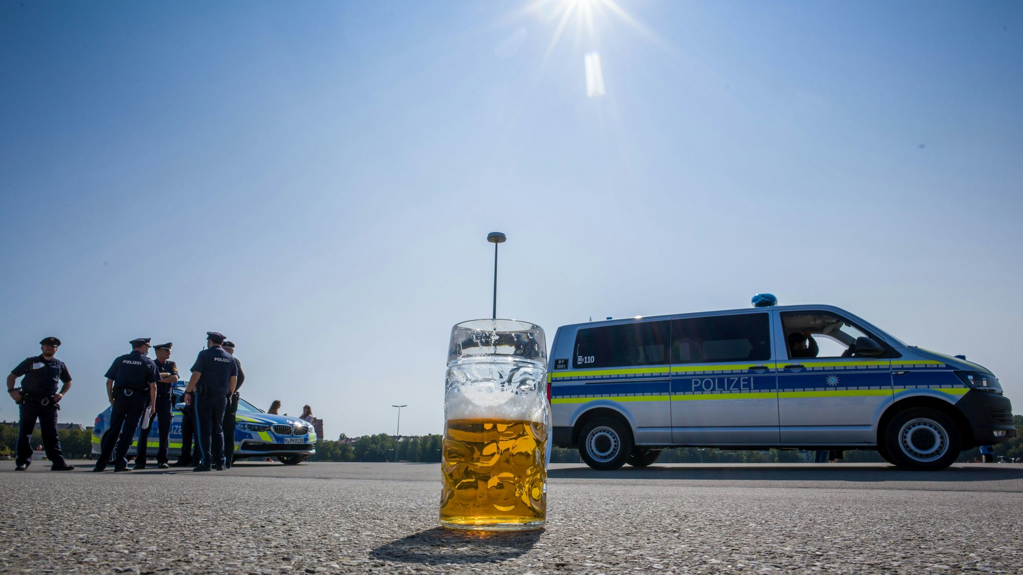 Ein verlassener, halb gefüllter Bierkrug auf dem Asphalt. Im Hintergrund stehen Polizisten neben Polizeifahrzeugen.