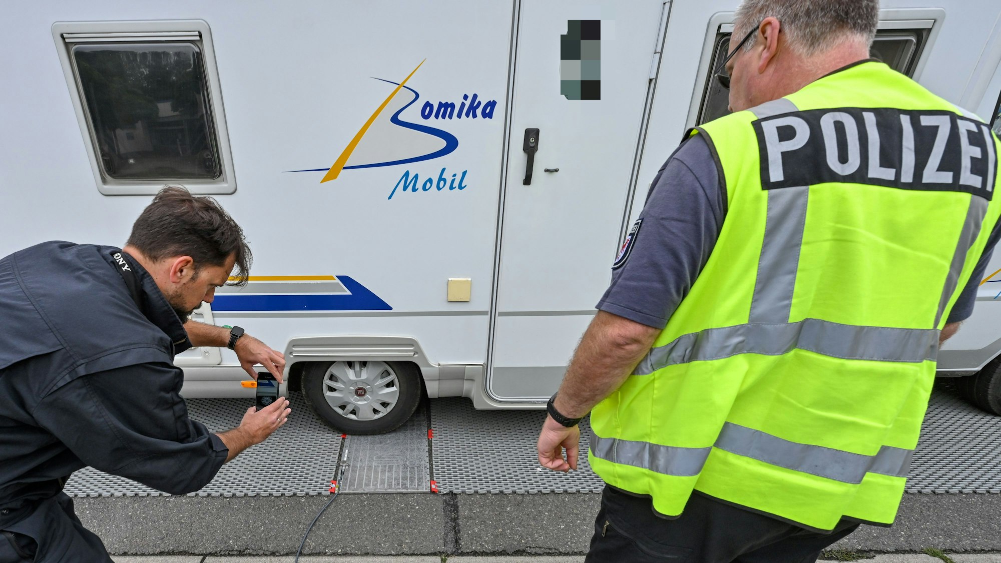 Ein Polizei steht neben einem Wohnmobil, das auf einer mobilen Waage steht. Ein zweiter Mann macht mit seinem Handy ein Foto.