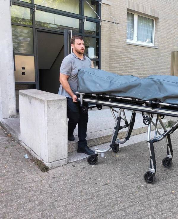 Zwei Bestatter bringen die Leiche auf einer Bahre mit Rollen aus dem Haus.