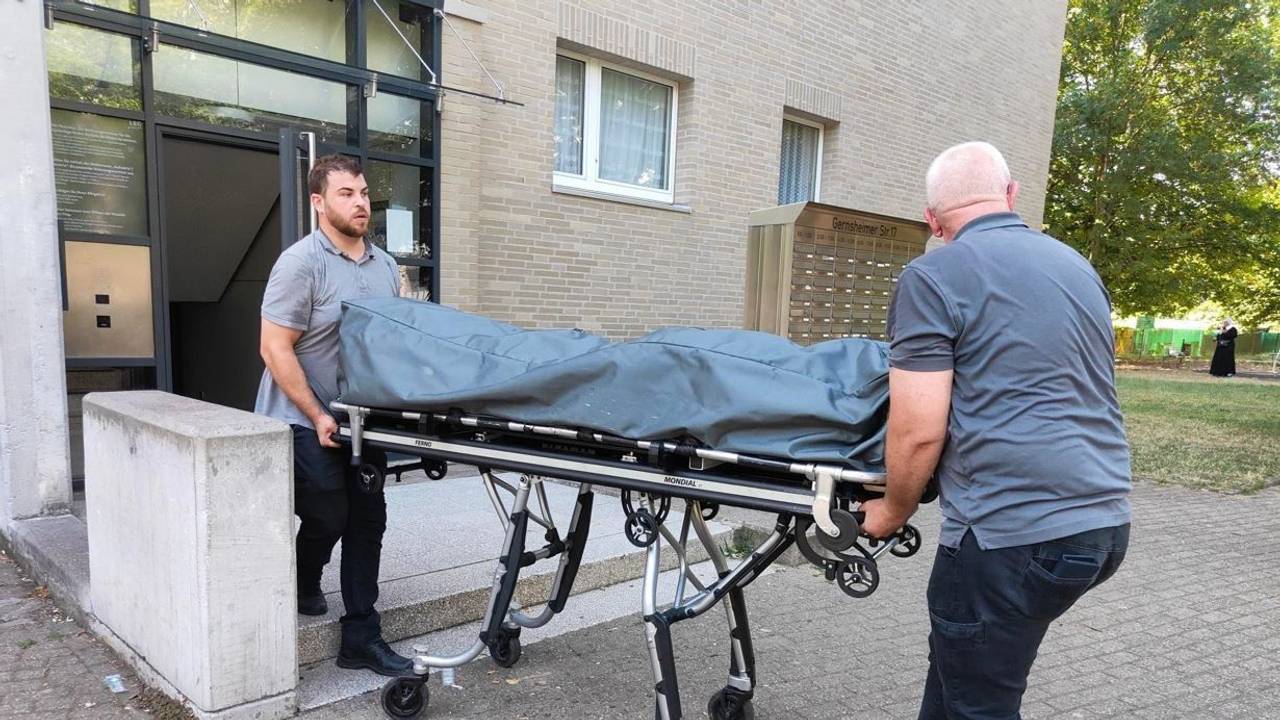 Zwei Bestatter bringen die Leiche auf einer Bahre mit Rollen aus dem Haus.