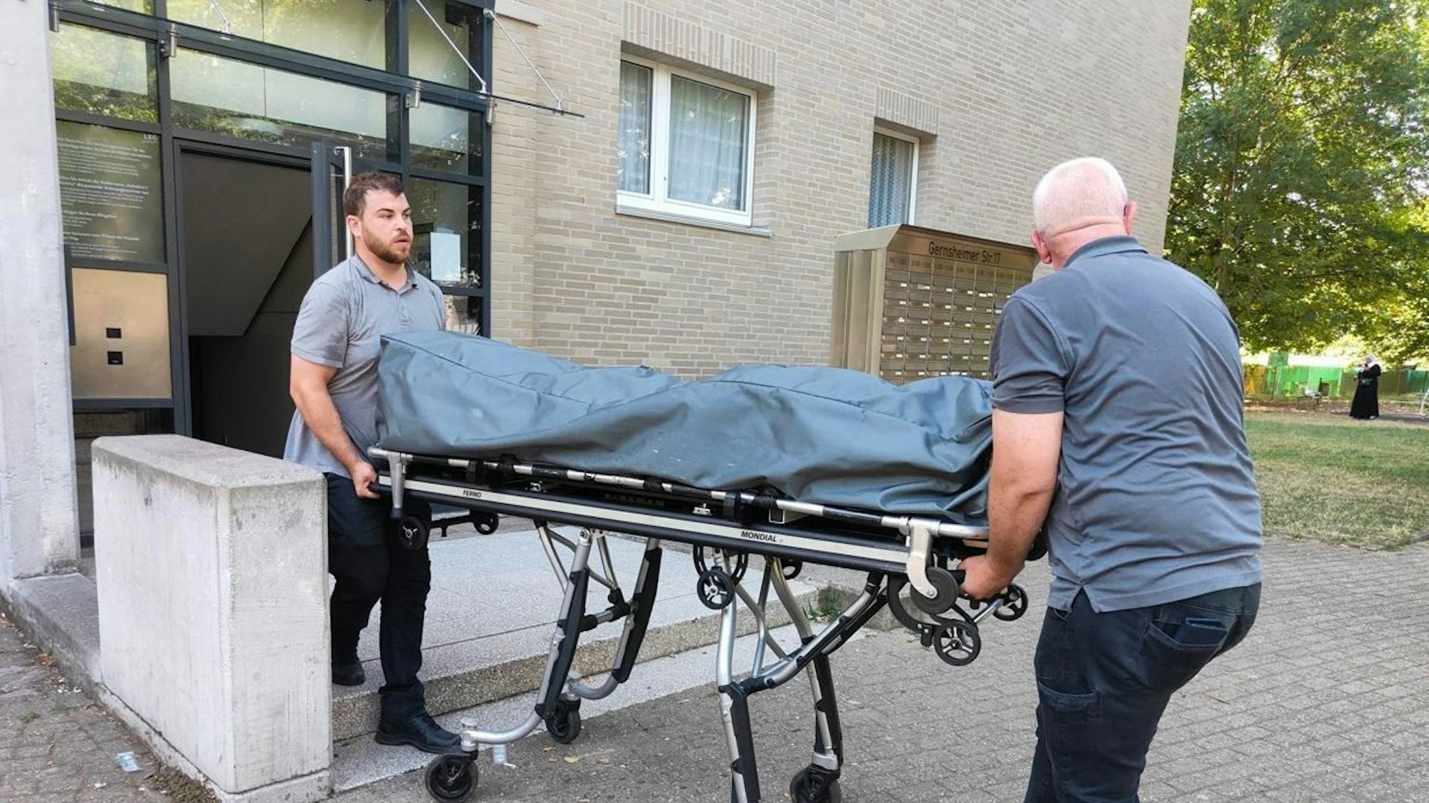 Zwei Bestatter bringen die Leiche auf einer Bahre mit Rollen aus dem Haus.