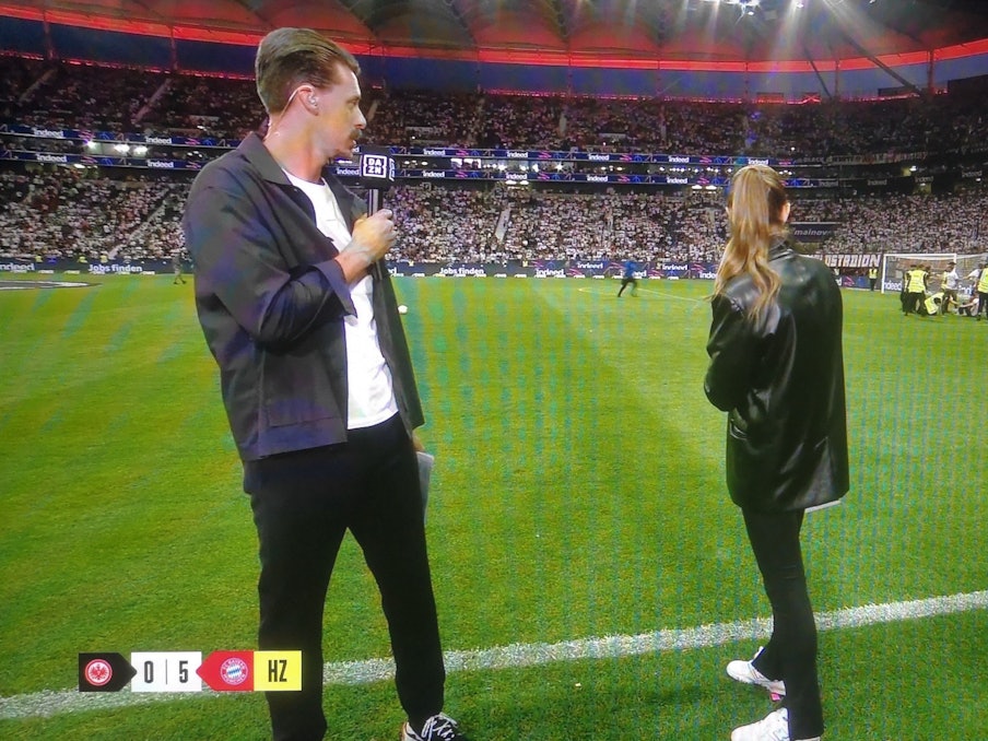 Laura Wontorra und Sandro Wagner blicken während der Halbzeit-Analyse bei DAZN auf den Rasen in Frankfurt, wo sich Fan-Chaoten einen Streit mit Ordnern liefern.