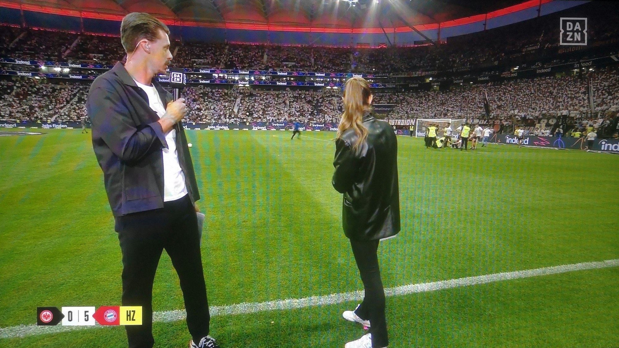 Laura Wontorra und Sandro Wagner blicken während der Halbzeit-Analyse bei DAZN auf den Rasen in Frankfurt, wo sich Fan-Chaoten einen Streit mit Ordnern liefern.