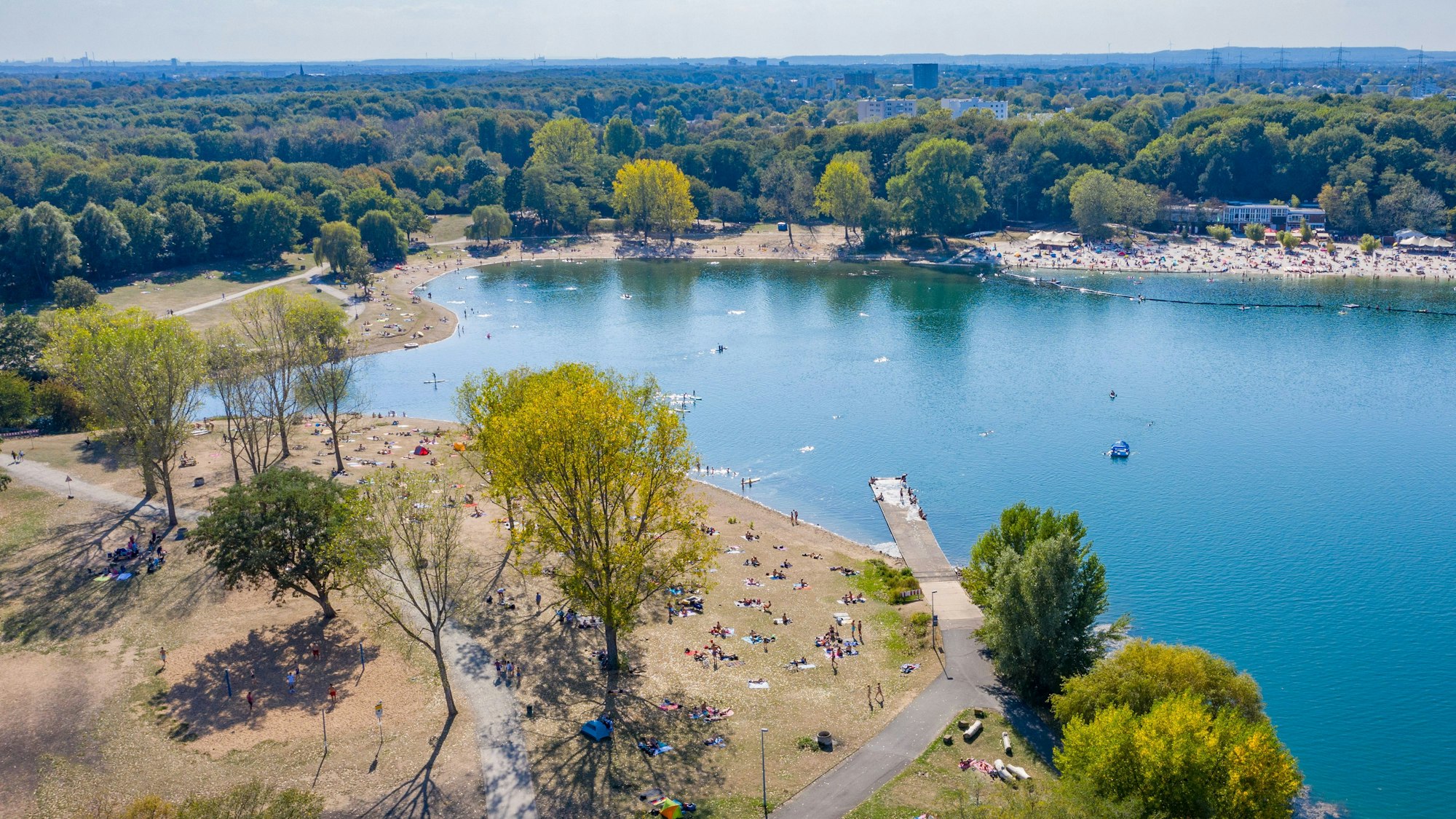 Der Fühlinger See von oben: An seinen Ufern liegen zahlreiche Menschen, ein Steg führt in den See, im Wasser sind vereinzelt Schwimmende.